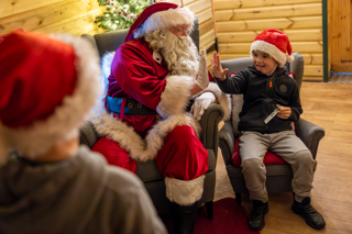 Santa Claus sits in a grey armchair inside a festive log cabin, giving a high-five to a child wearing a Santa hat who is sitting in a smaller chair beside him.