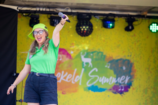 A smiling woman wearing a green polo shirt, black shorts, glasses and a neon-green cap holds up a microphone on a stage against a colourful "Stockeld Summer" backdrop.