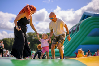 Two adults help a smiling toddler walk on an inflatable play area outdoors, with other children playing and large slides visible in the background.