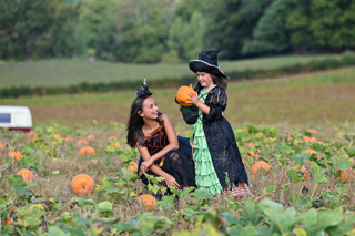 A young girl dressed in a witch’s costume stands in a pumpkin patch holding a pumpkin whilst another girl in fancy dress kneels beside her. 