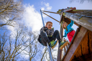 Two children enjoying a sunny day at a playground. One slides down a pole while the other watches from a wooden platform. 