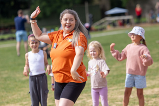 A woman in an orange shirt leads an outdoor dance with children on a sunny day. 