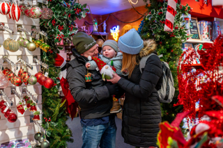 Two adults, carrying a young child, smile while holding a holiday decoration inside Stockeld Park’s Christmas shop, filled with colourful hanging ornaments.