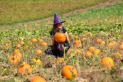 girl in purple witches hat holding an orange pumpkin in a pumpkin field