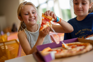 Two young girls sit together with a pizza box laid in front of them, whilst one pulls a cheesy slice from the box. 