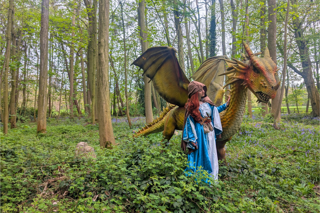A woman dressed as a medieval storybook character standing next to a dragon statue in a lush green forest.