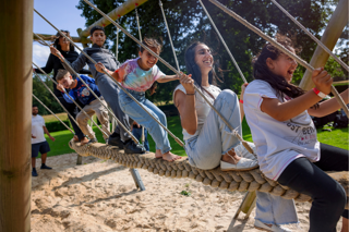  A cheerful group of children, teenagers and adults playing together on a swinging rope bridge over sand at an outdoor playground.