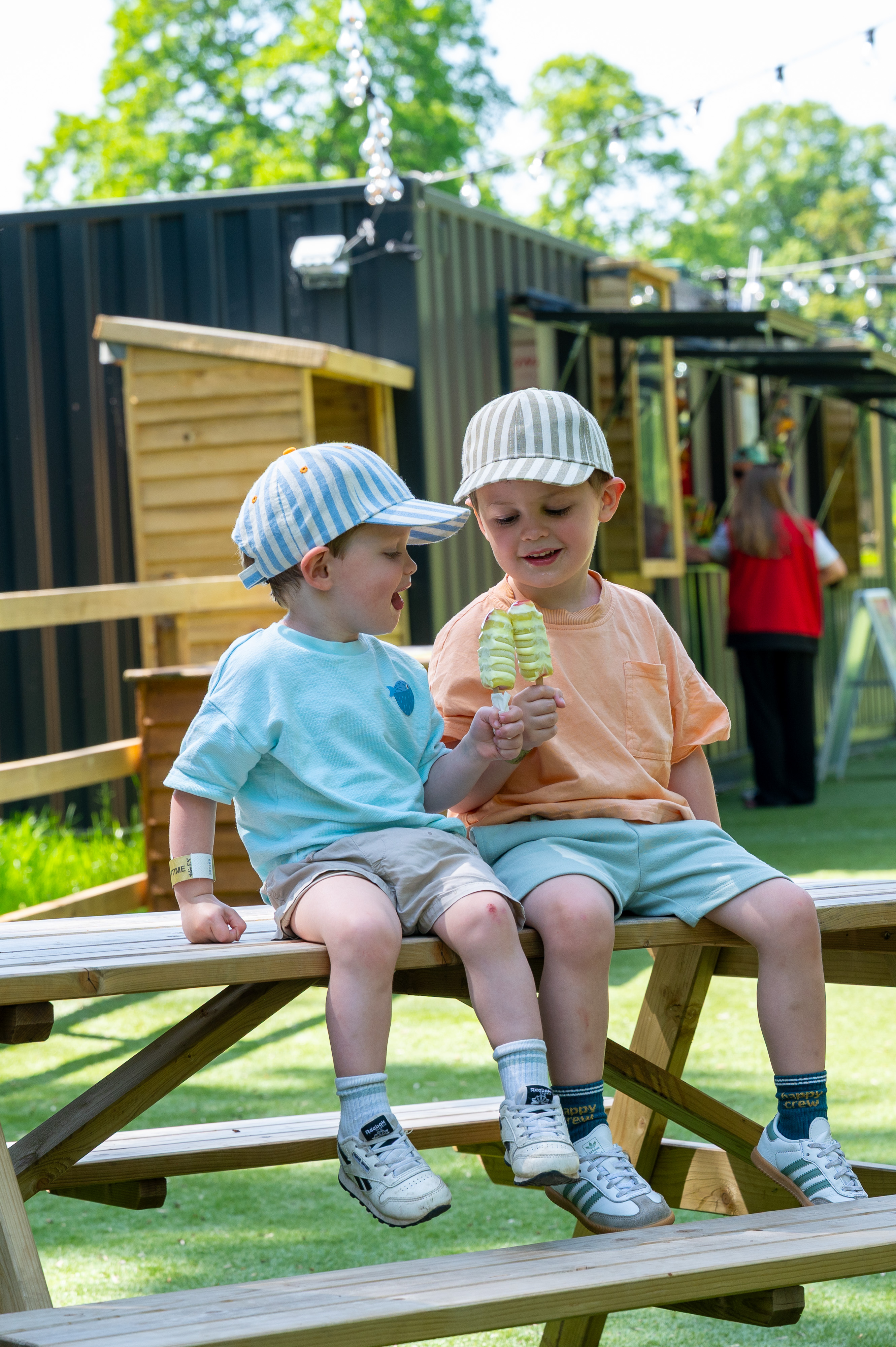 two young guests sitting on a picnic bench wearing stripey hats with ice lollies