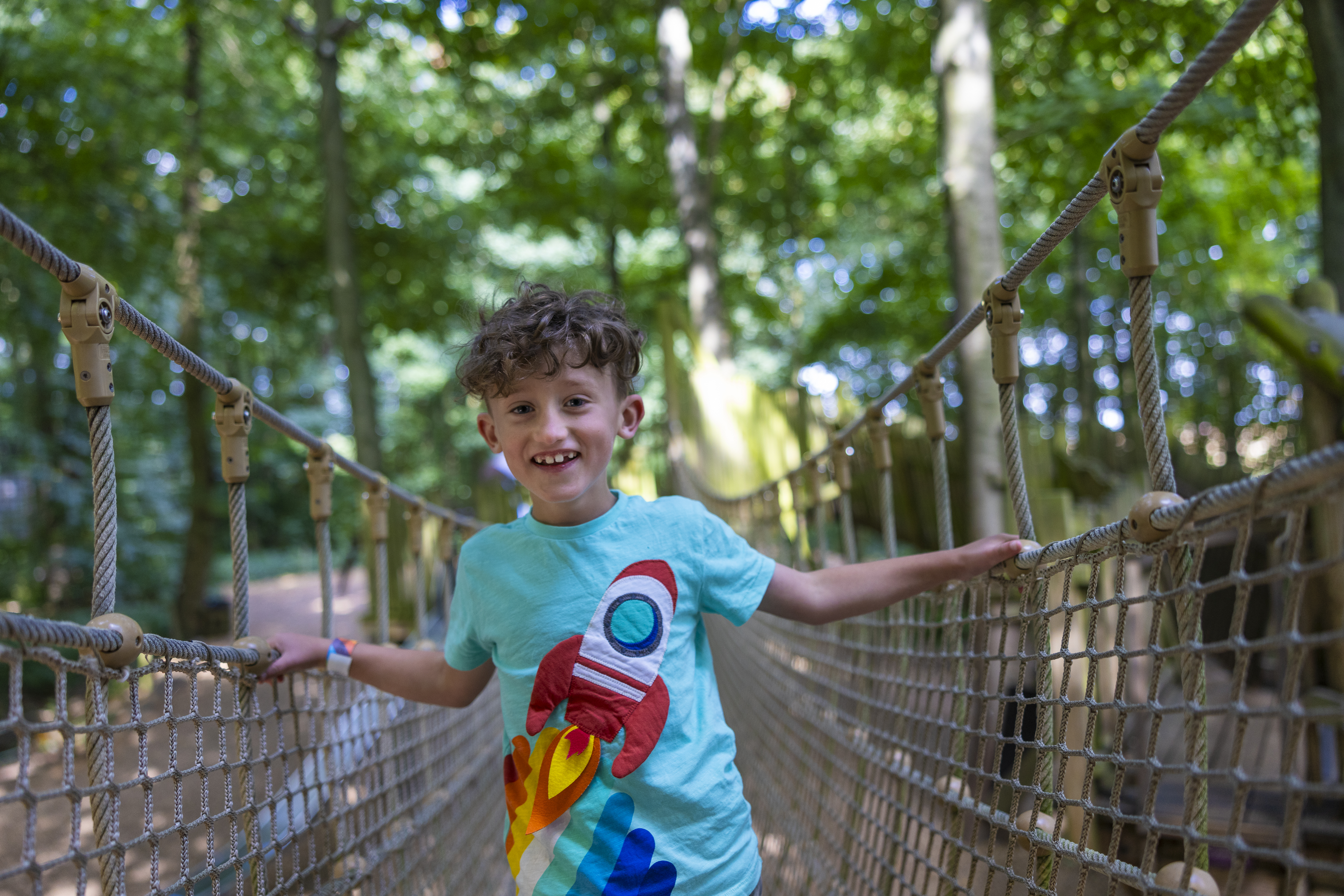 boy wearing blue tshirt with a rocket on it standing on a rope bridge in a forest