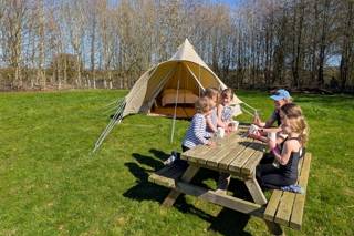 A group of children and an adult sit around a wooden picnic table, drinking from mugs on a sunny day in a grassy field next to a large bell tent.