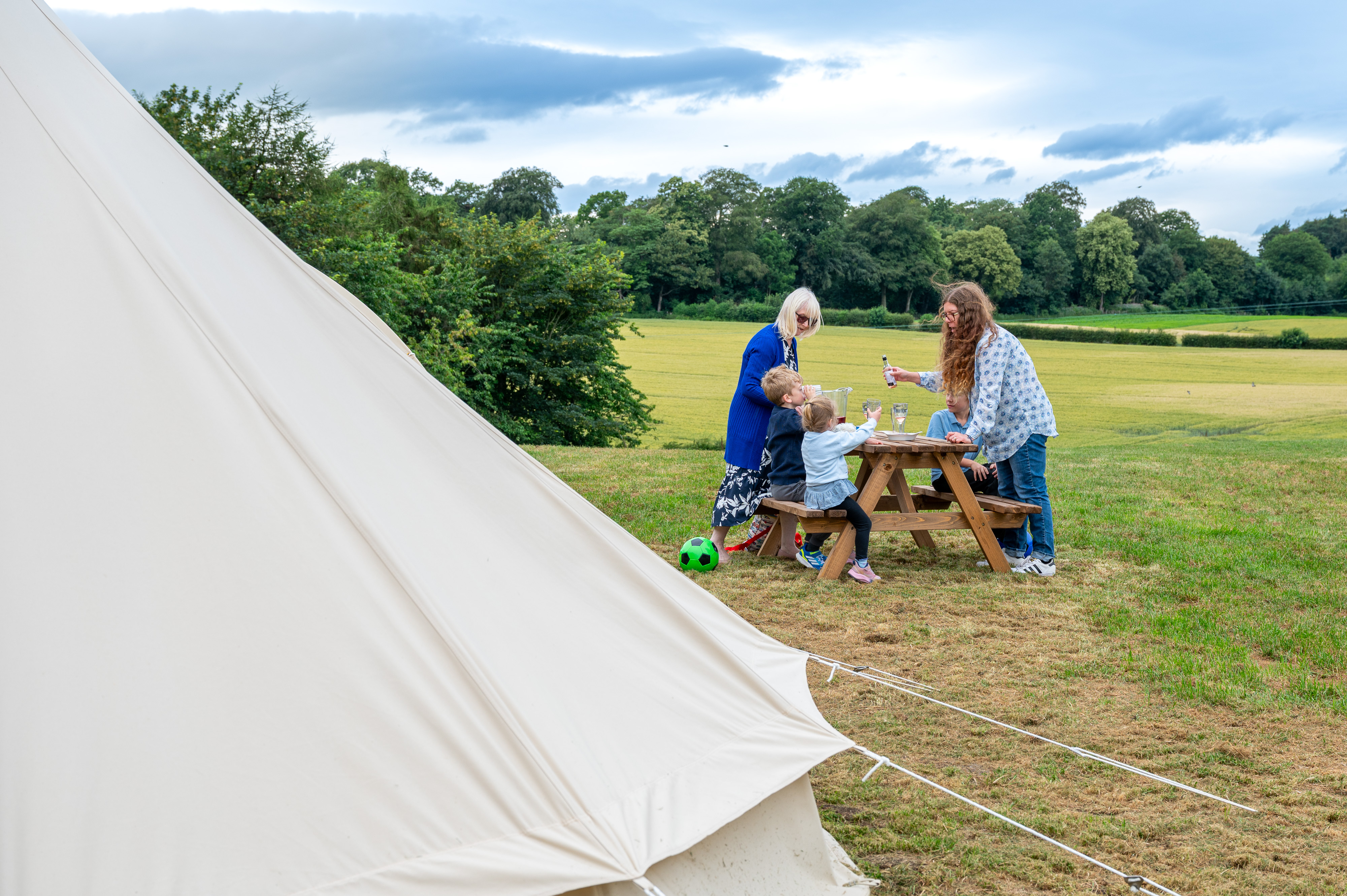 Family Sat At A Picnic Bench In Front Of A Bell Tent With Fields