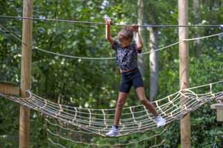 girl in black tshirt and shorts climbing across net while holding rope in a forest