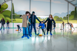 A group of people ice skating together on a covered indoor rink, with one person using a penguin-shaped skating aid for balance.