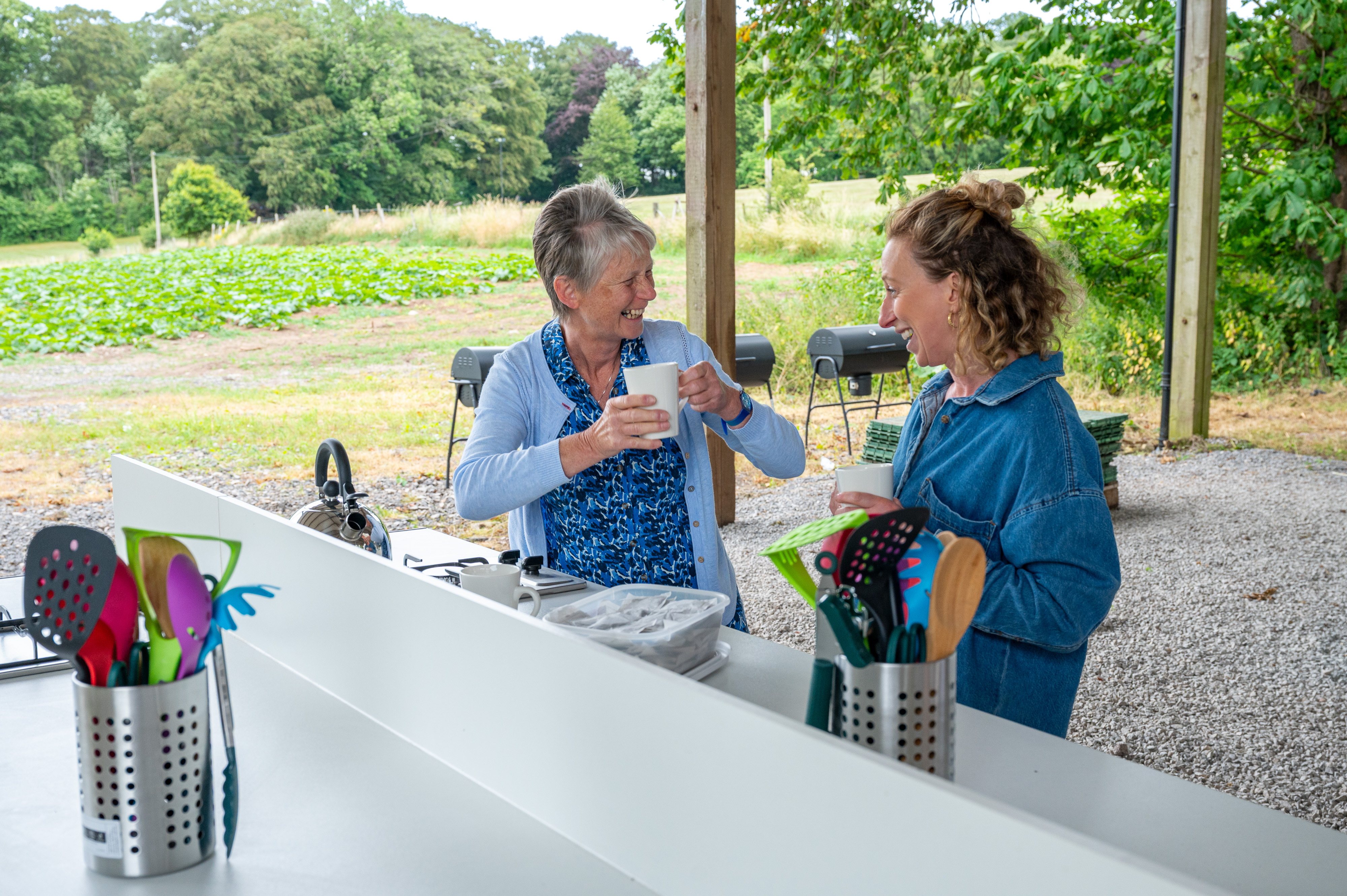 Two Women Laughing While Drinking Cup Of Tea In Outdoor Kitchen