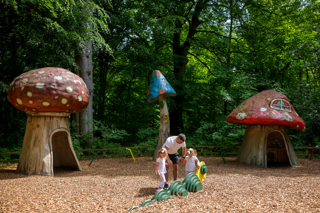 A man and two young girls play on a playground in a forest, surrounded by large playhouses shaped like colourful fairytale mushrooms.