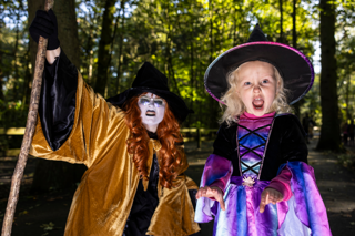 Woman dressed in a witch costume crouches next to a young girl dressed in a witch costume outside in a fore