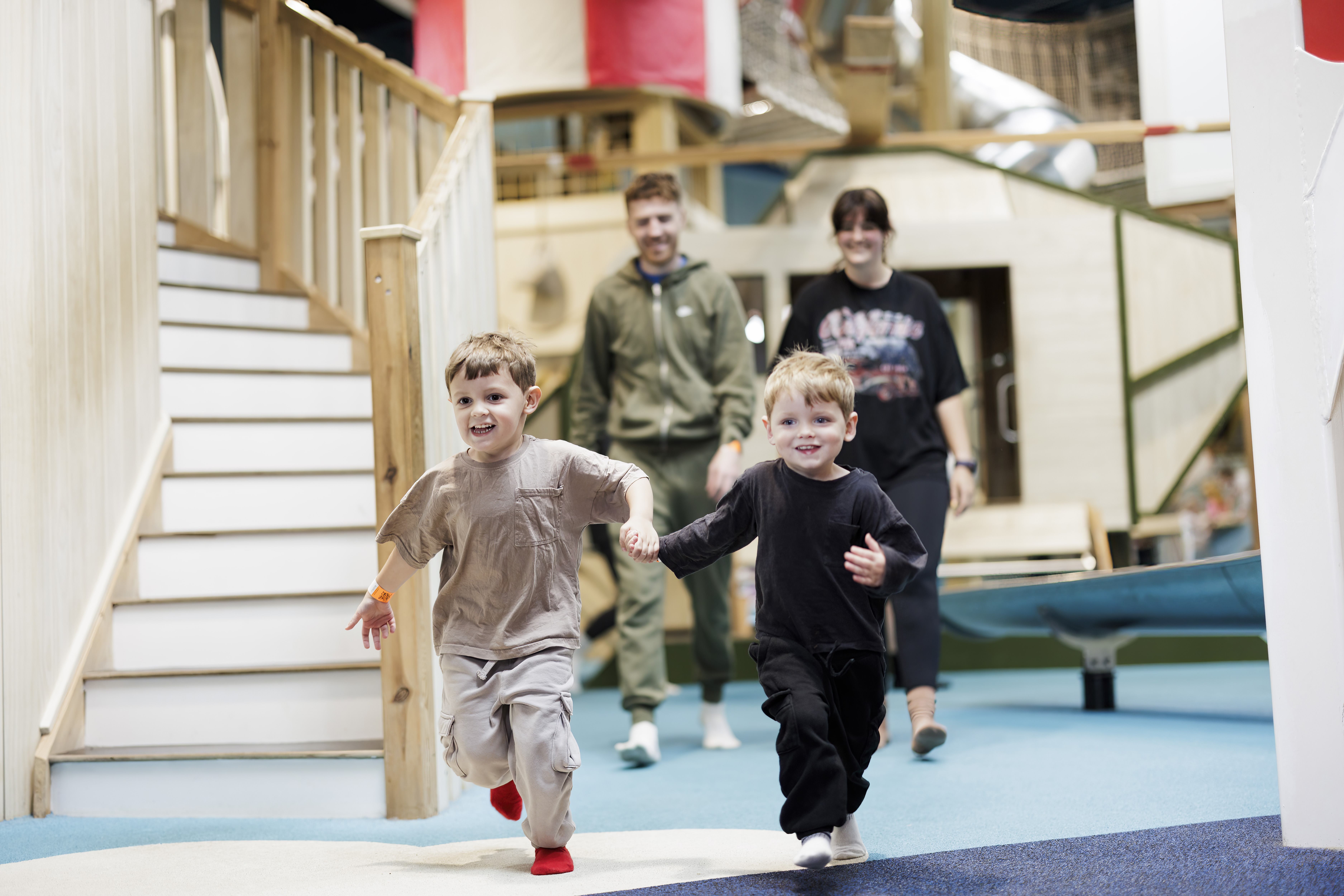 two young guests running holding hands through indoor play area with two parents walking behind smiling