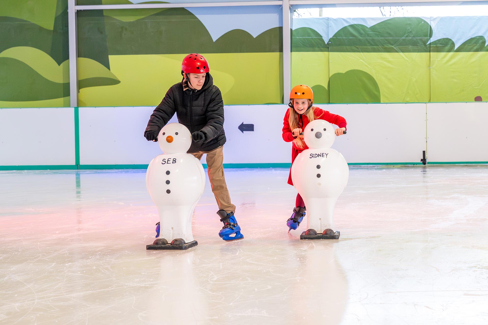 brother and sister ice skating together in covered rink with snowmen skating aids