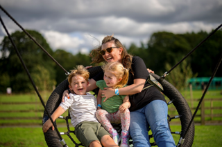 An adult and two young children sit together on a large round swing outdoors with a field and trees in the background.