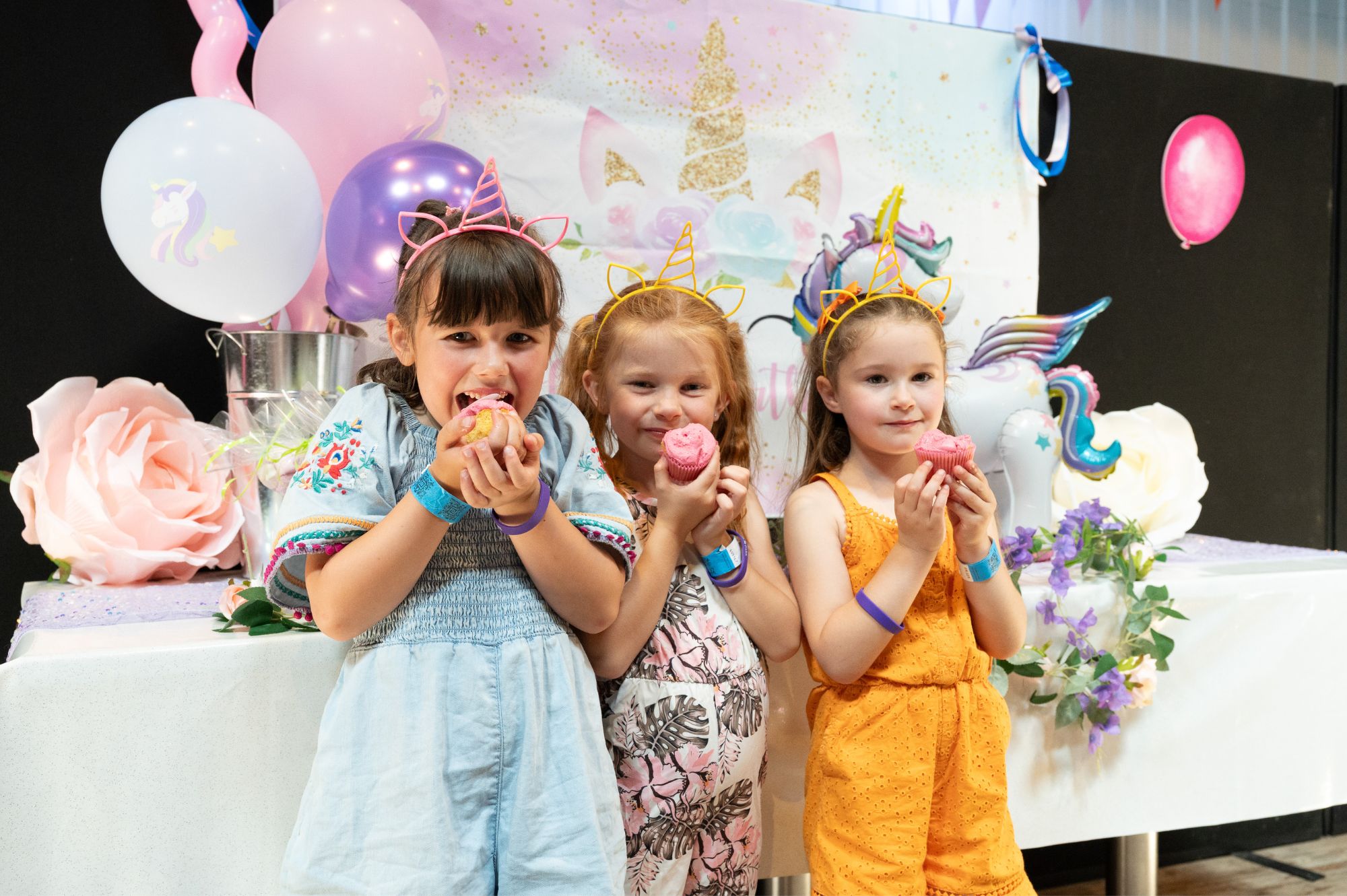 Three young girls eat cupcakes and wear unicorn themed party headbands.