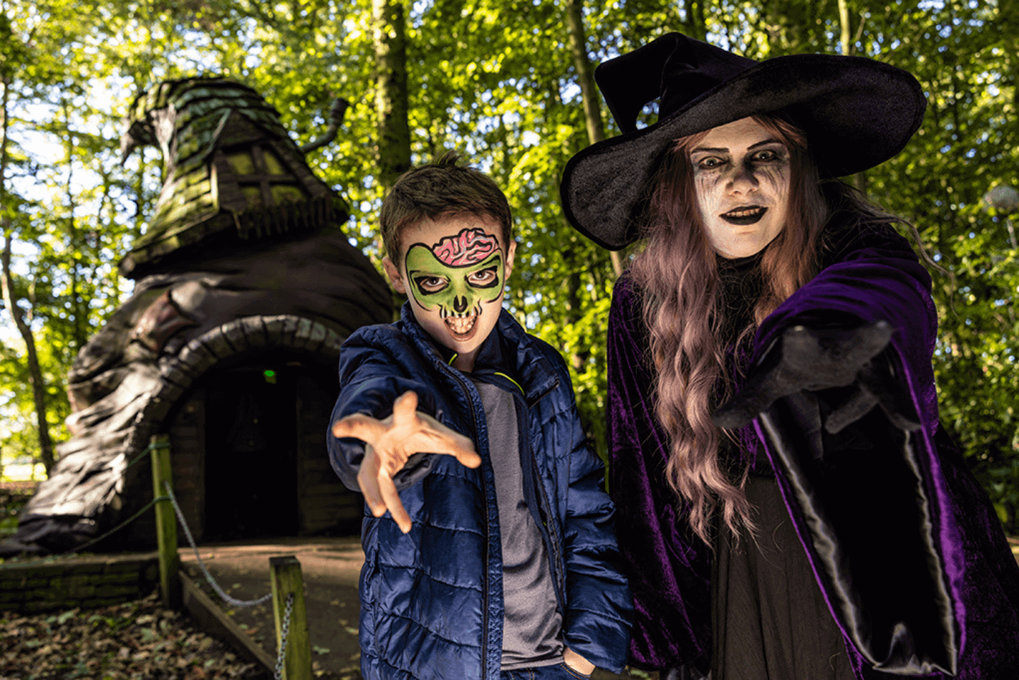 Young child with face paint stands next to a woman dressed as a witch, situated in a forest 